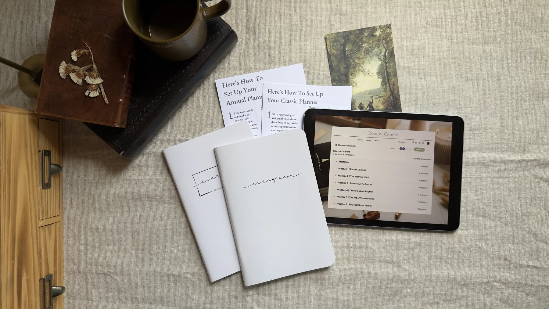 The Getting Started Kit laid out on a desk: two Evergreen planner booklets, two printed setup guides, a mug, a leather journal, and an iPad playing the Renew course.