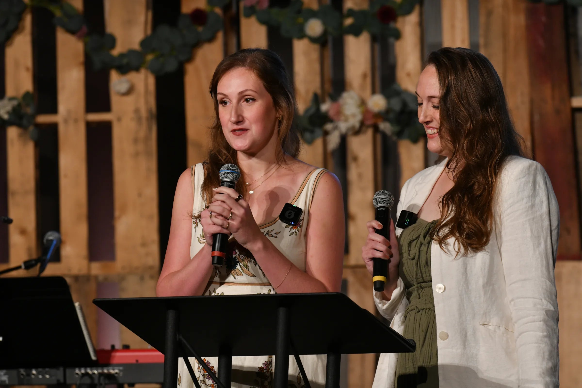 Two women teaching from a stage, each holding a microphone behind a lectern, in front of a wood-panel backdrop hung with eucalyptus and flowers.